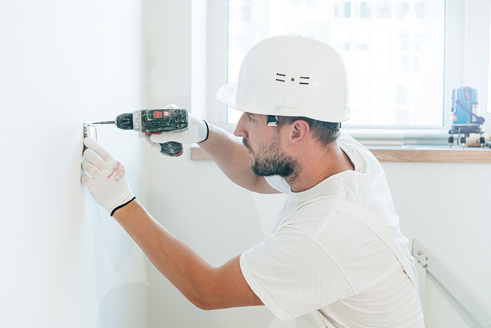 Fit-out technician in a white hard hat and overalls drilling into a wall in a bright room