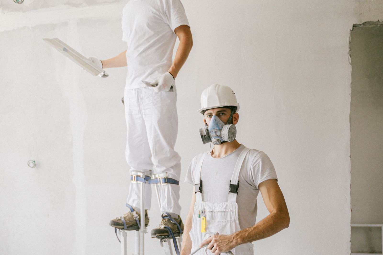 Two painters in white overalls and respirators preparing a wall, one working from stilts