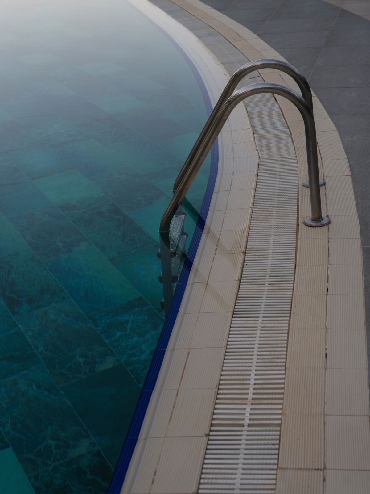 Swimming pool edge with a metal ladder and clear water over blue tiles