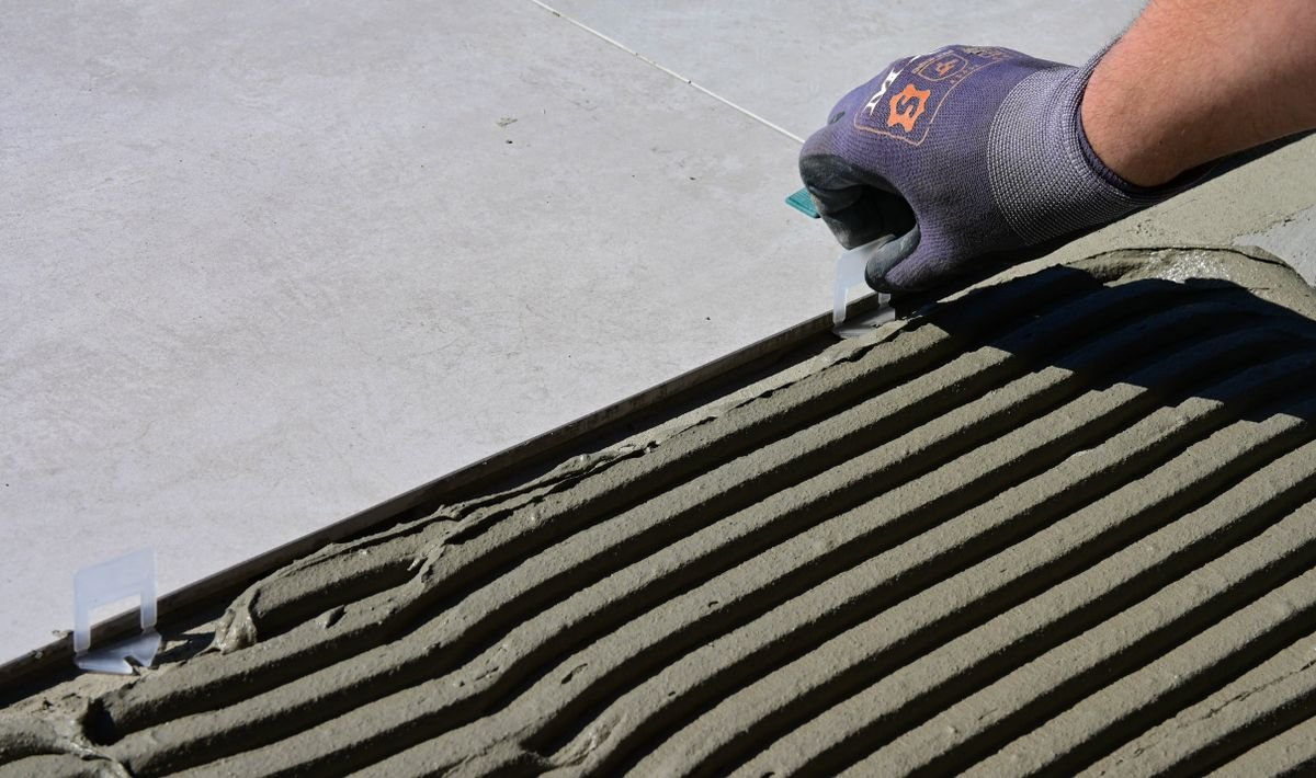 Notched-trowel adhesive ridges beside a large porcelain tile, with a gloved hand at the edge