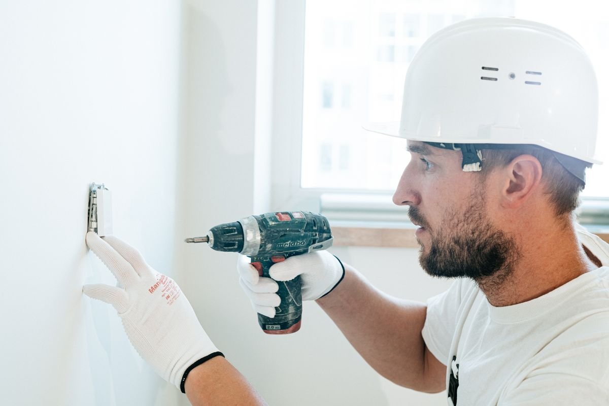 Technician in a hard hat drilling a fixing into a wall beside a window